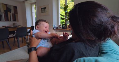 Anne, anne ve çocuk arasındaki bağ ve sevgi dolu doğal ışıkla dolu parlak bir oturma odasında oynarken, gülümserken ve gülerken bebeği havaya kaldırıyor.