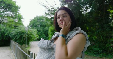 Bir taşıyıcıda bebeğini taşıyan anne, parkta dikilirken elini yüzünü dinlendirirken, bir anlık derin düşüncelere dalmış ve çevresiyle olan bağlantısını yansıtıyor.