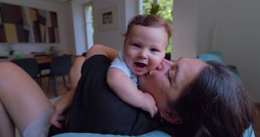 Anne sıcak ve sevgi dolu bir anda bebeği öpüyor ve kucaklıyor, doğal ışık sıcak bir oturma odasına akıyor, anne ve çocuk arasında neşeli ve samimi bir bağ kuruyor.