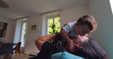 Anne, anne ve çocuk arasındaki bağ ve sevgi dolu doğal ışıkla dolu parlak bir oturma odasında oynarken, gülümserken ve gülerken bebeği havaya kaldırıyor.