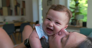 Anne, sevgi dolu ve sevgi dolu bir anda bebeği öpüyor ve kucaklıyor. Parlak bir oturma odasında oturuyor. Neşe ve yakınlık içinde kaynaşıyor.