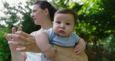 Yeşil bir parkta annesi tarafından tutulan bebek, şaşkın ve meraklı bir ifadeyle, doğal bir çevrede keşif ve keşif anını yakalıyor.