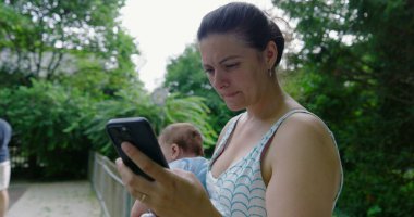 Anne akıllı telefonunu açık havada kullanırken bebeği kucağına alıyor, sonra da kaldırıyor, çocuğuyla yeşil bir park ortamında etkileşime geçiyor.