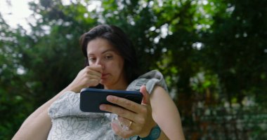 Anne bebeğini bir taşıyıcıda tutarken burnunu kaşıyor, akıllı telefonuyla sakin bir park ortamında meşgul, modern ebeveynliği, teknolojiyi ve doğal çevreyi harmanlıyor.