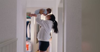 Anne sevinçle bebeğini havaya kaldırıyor, aydınlık bir koridorda sevgi dolu bir anı paylaşıyor. neşeli etkileşimler ve gülümsemeler anne ve çocuk arasındaki güçlü bağı ve mutluluğu gösterir..