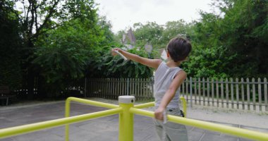 Çocuk sarı bir atlıkarıncada dikilirken elinde sopayla arka planda yeşillik ve çitle