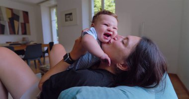 Anne sıcak ve sevgi dolu bir anda bebeği öpüyor ve kucaklıyor, doğal ışık sıcak bir oturma odasına akıyor, anne ve çocuk arasında neşeli ve samimi bir bağ kuruyor.