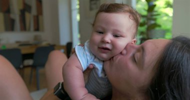 Anne, sevgi dolu ve sevgi dolu bir anda bebeği öpüyor ve kucaklıyor. Parlak bir oturma odasında oturuyor. Neşe ve yakınlık içinde kaynaşıyor..