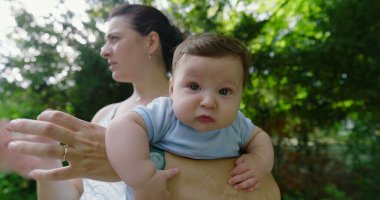 Yeşil bir parkta annesi tarafından tutulan bebek, şaşkın ve meraklı bir ifadeyle, doğal bir çevrede keşif ve keşif anını yakalıyor..