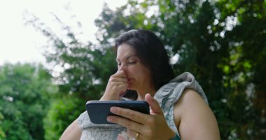 Anne bebeğini bir taşıyıcıda tutarken burnunu kaşıyor, akıllı telefonuyla sakin bir park ortamında meşgul, modern ebeveynliği, teknolojiyi ve doğal çevreyi harmanlıyor..