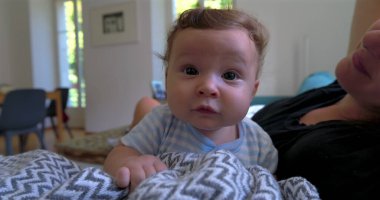 Bebek battaniyeye uzanmış merakla bakarken anne arka planda doğal ışık ve sıcak aile atmosferiyle dolu rahat bir oturma odasında gülümsüyor.