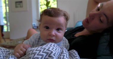 Bebek battaniyeye uzanmış merakla bakarken anne arka planda doğal ışık ve sıcak aile atmosferiyle dolu rahat bir oturma odasında gülümsüyor.
