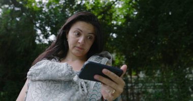 Anne endişeli bir ifadeyle, bebeğini bir taşıyıcıda tutarken dikkatle parktaki akıllı telefonuna odaklanıyor, modern endişelerin ve ebeveynliğin karışımını vurguluyor.
