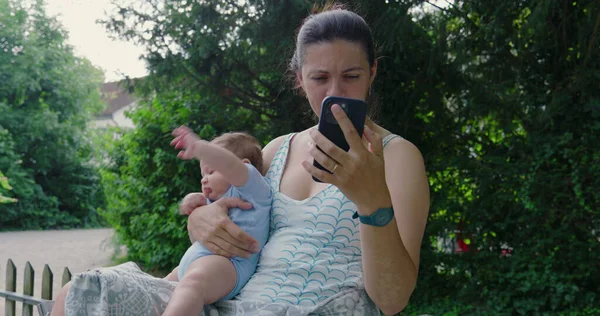 Anne akıllı telefonuyla bir video görüşmesi yaparken bebeği kucağında tutuyor, anne yaz günü sohbete odaklanıyor.