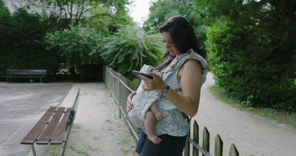 Anne endişeli bir ifadeyle, bebeğini bir taşıyıcıda tutarken dikkatle parktaki akıllı telefonuna odaklanıyor, modern endişelerin ve ebeveynliğin karışımını vurguluyor.