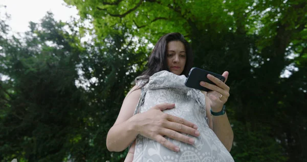 Parkta çoklu görev yapan bir kadın, akıllı telefonuna odaklanırken bebeğini bir taşıyıcıda tutuyor, ağaçlarla çevrili huzurlu bir açık hava ortamının tadını çıkarıyor..