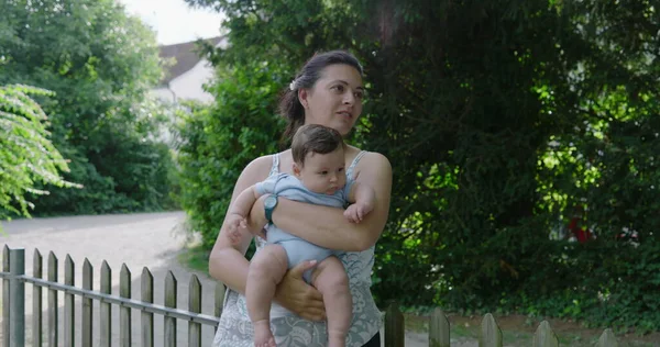 Anne dışarıda bebeğini tutuyor, çocuğuyla meşgul olurken gülümsüyor, arka planda yeşillikler ve ağaçlar var, doğal park ortamında neşeli ve hassas bir an.