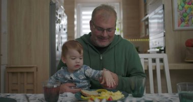 Babam yemek masasında kucağında bebeğini tutarken gülümsüyor, sıcak ve sevgi dolu bir aile anı yaratıyor.