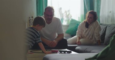 Baba kahve masasında oyuncak trenle oynayan çocuğu izliyor, anne arka planda küçük beyaz bir köpek tutuyor, aile bağı ve sakin bir oturma odasında oyun zamanı.