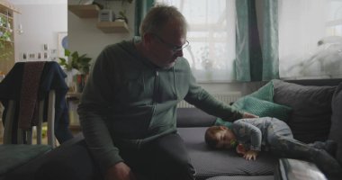 Babam, bir bebeğin yanında, emzik ile bir kanepede dinleniyor. Huzurlu bir aile salonunda, içi doğal ışıkla dolu bir el uzatıyordu.