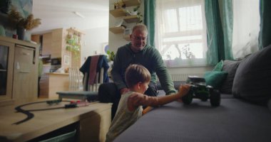 Babası gülümserken oğlu kanepede yeşil bir oyuncak kamyonla oynuyor. Sıcak ve rahat bir oturma odasında, ahşap mobilyaları ve canlı bir iç mekanı var.