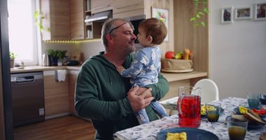 Baba kucağında bebek ve gülümsüyor. Sıcak bir mutfak ortamında neşeli ve sevecen bir an yaşıyor.