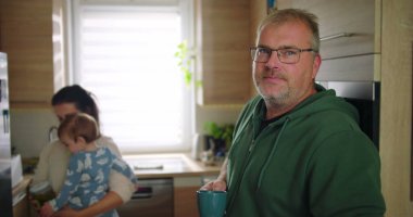 Annesi buzdolabını kollarında bebekle kontrol ederken elinde bardak tutan bir adam, tipik bir sabah rutini sırasında sıradan bir aile anı, sıcak ve besleyici ebeveynlik sahnesi.