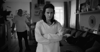 Tartışma sırasında kollarını çapraz uzatmış bir kadın, arka planda yürüyen ve el kol hareketi yapan bir adam, gergin ve duygusal ilişki çatışması, siyah ve beyaz