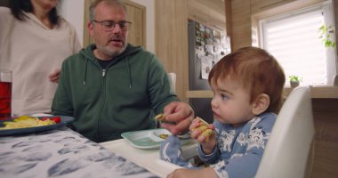 Baba yemek sırasında bebek maşasında oturur, anne tabağın yanında durur, aile bağlarını yemek masasında kurar, çocuk yemek yer.
