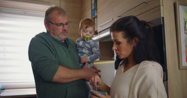 Anne modern mutfakta kahvesini karıştırırken baba kucağında. Ev ortamında sakin bir aile ortamının altını çiziyor.