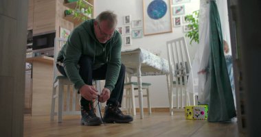 Babam, sıcak ahşap döşemeleri ve doğal ışıklandırması olan sıcak bir aile yemek odasında, emzik ile perdelerin yanında duran bir çocuğa bakarken botlarını bağlıyor.