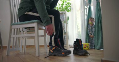 Babam bir sandalyede oturmuş botlarını bağlarken bir bebek elinde renkli bir oyuncak bloğuyla perdelerin yanında duruyor. Ahşap zemin ve doğal ışık olan bir aile yemek odasında.