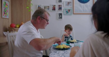 Baba ve küçük oğlu mutfakta öğle yemeği yiyorlar, sıradan aile anları, yemek servisi yapan babaya odaklan, çocuk dikkatle dinliyor, rahat bir atmosfer.