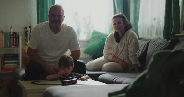 Anne ve baba kanepede oturup çocuğa gülümsüyor kahve masasında oyuncak trenle oynuyor rahat oturma odası düzeninde, neşeli aile anları, kapalı alanda kaynaşma zamanı.