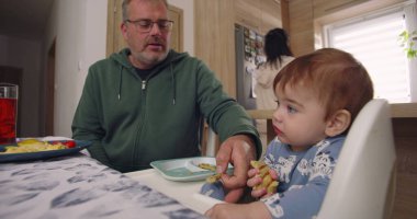 Baba yemek sırasında bebek maşasında oturur, anne tabağın yanında durur, aile bağlarını yemek masasında kurar, çocuk yemek yer.