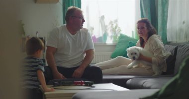 Baba ve çocuk sehpanın üzerinde oyuncak trenle etkileşime giriyor, anne arka planda beyaz köpeği tutuyor, yakın aile bağı ve kapalı alanda kaynaşma.