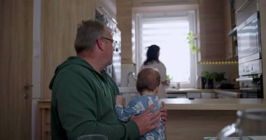 Baba bebeğini kucağında tutarken anne mutfakta hazırlanıyor sıcak ve sevgi dolu bir aile ortamında ev ortamında.