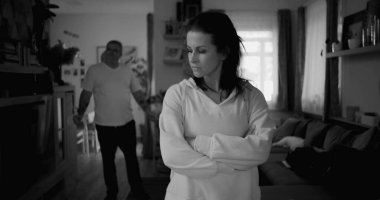 Tartışma sırasında kollarını çapraz uzatmış bir kadın, arka planda yürüyen ve el kol hareketi yapan bir adam, gergin ve duygusal ilişki çatışması, siyah ve beyaz