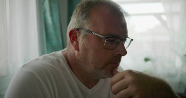 Adamın biri, çenesini elinde dinlendirirken, sakin ve rahat bir evde yumuşak yeşil perdeleri olan bir pencerenin yanında otururken düşünceli bir şekilde bakıyor.