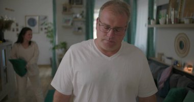 Oturma odasında hararetli tartışmalar yaşayan bir çift, görünürde üzgün duran bir adam, arka planda duygusal hareketler yapan bir kadın, ilişki çatışması sırasında gergin bir an.