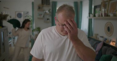 Oturma odasında hararetli tartışmalar yaşayan bir çift, görünürde üzgün duran bir adam, arka planda duygusal hareketler yapan bir kadın, ilişki çatışması sırasında gergin bir an.