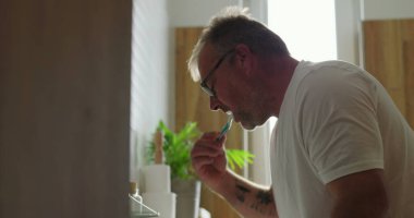 Banyo lavabosunda dişlerini fırçalayan adam, yoğun ifadeli yan profil görüntüsü, parlak ve temiz ev ortamında sabah diş bakımı rutini