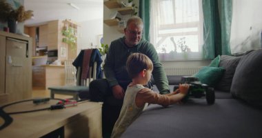 Babası gülümserken oğlu kanepede yeşil bir oyuncak kamyonla oynuyor. Sıcak ve rahat bir oturma odasında, ahşap mobilyaları ve canlı bir iç mekanı var.