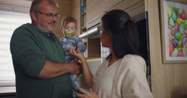 Anne modern mutfakta kahvesini karıştırırken baba kucağında. Ev ortamında sakin bir aile ortamının altını çiziyor.