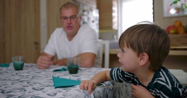 Erkek ve çocuk masada sakin bir anı paylaşırken, bir kadın arka planda çalışıyor, samimi bir evde samimi bir aile etkileşimi sergiliyor.