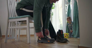 Babam bir sandalyede oturmuş botlarını bağlarken bir bebek elinde renkli bir oyuncak bloğuyla perdelerin yanında duruyor. Ahşap zemin ve doğal ışık olan bir aile yemek odasında.