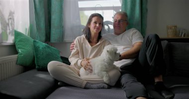 Küçük beyaz köpekle kanepede dinlenen, sakin ve samimi aile anlarının tadını çıkaran, doğal ışık ve yumuşak mobilyaları olan rahat oturma odası, sıcak atmosferi olan çift.