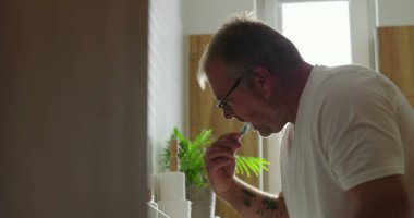 Banyo lavabosunda dişlerini fırçalayan adam, yoğun ifadeli yan profil görüntüsü, parlak ve temiz ev ortamında sabah diş bakımı rutini