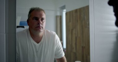 Profil görünümlü bir adam banyoda öne doğru eğiliyor, derin düşüncelere dalmış, içi beyaz fayanslarla dolu bir banyo ve ahşap kaplama, sessiz ve yansıtıcı kişisel bir an.