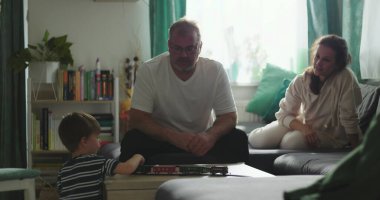 Babam, kahve masasında oyuncak trenle oynayan bir çocukla oturuyor. Anne, kanepeden gülümsüyor ve gözlemliyor. Sıcak aile bağları ve huzurlu ev ortamında samimi bir bağ.